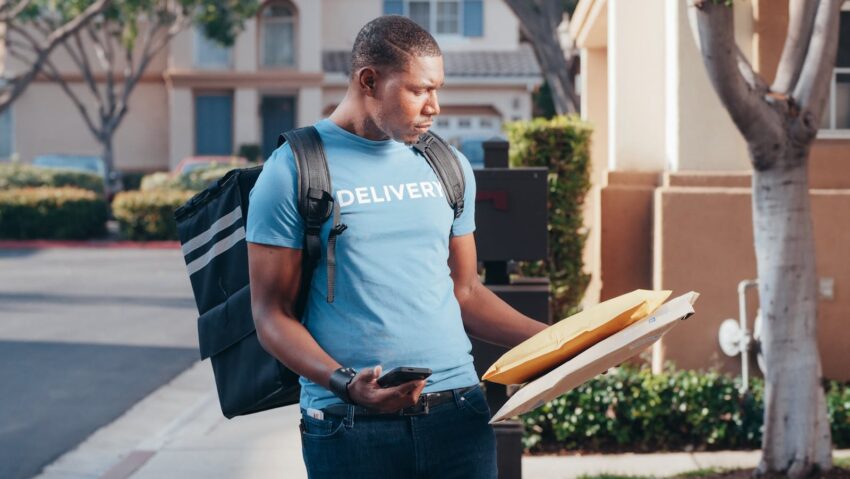 Delivery person with smartphone and parcels outside a house in a sunny neighborhood.